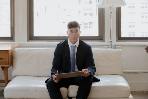 Free Young businessman in a suit sitting with a clipboard in a bright office setting. Ideal for corporate themes. Stock Photo