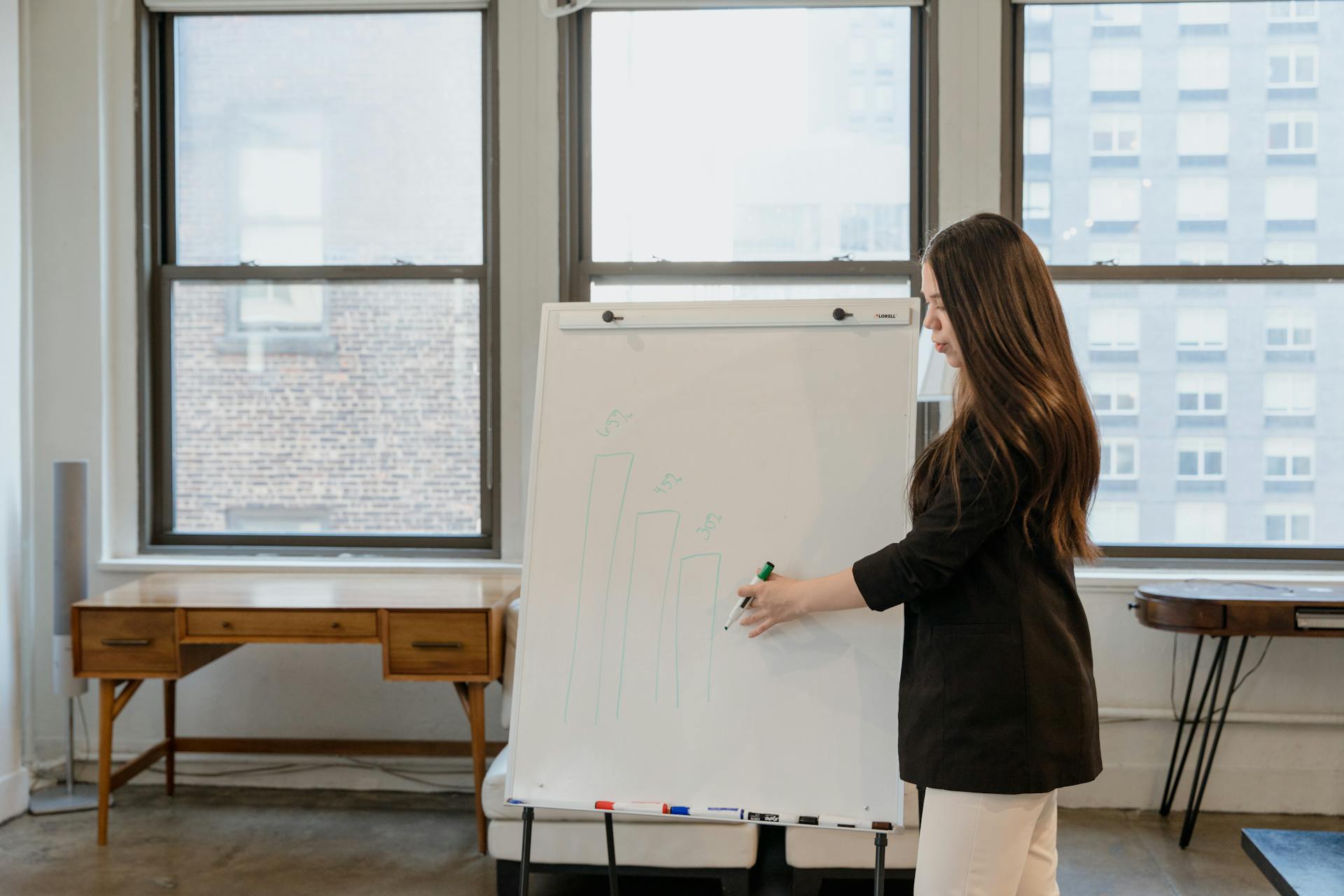 Businesswoman in black blazer presenting on a whiteboard with financial graphs in a modern office.