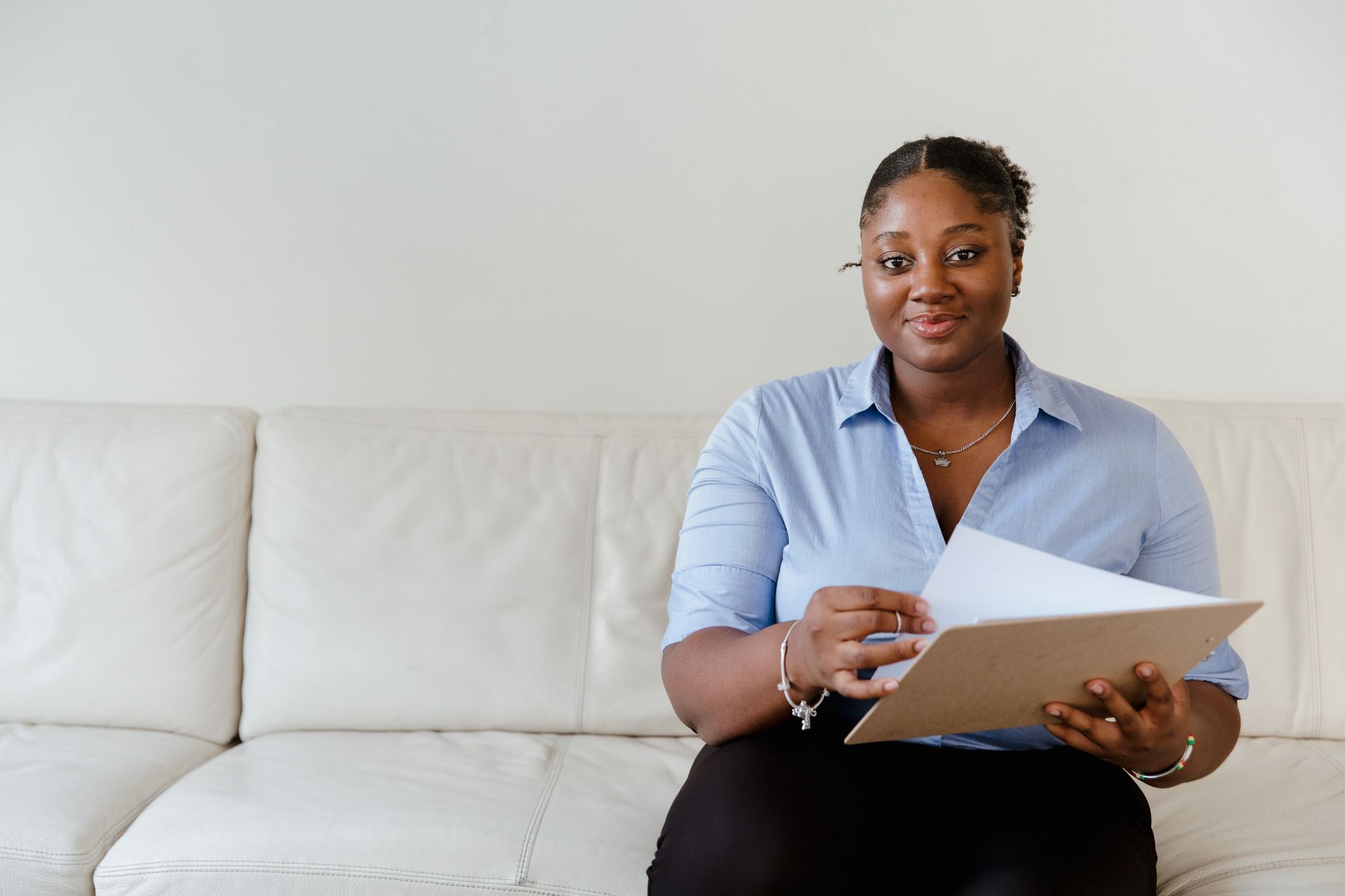A confident woman in blue shirt sitting on a white sofa holding a clipboard, smiling.