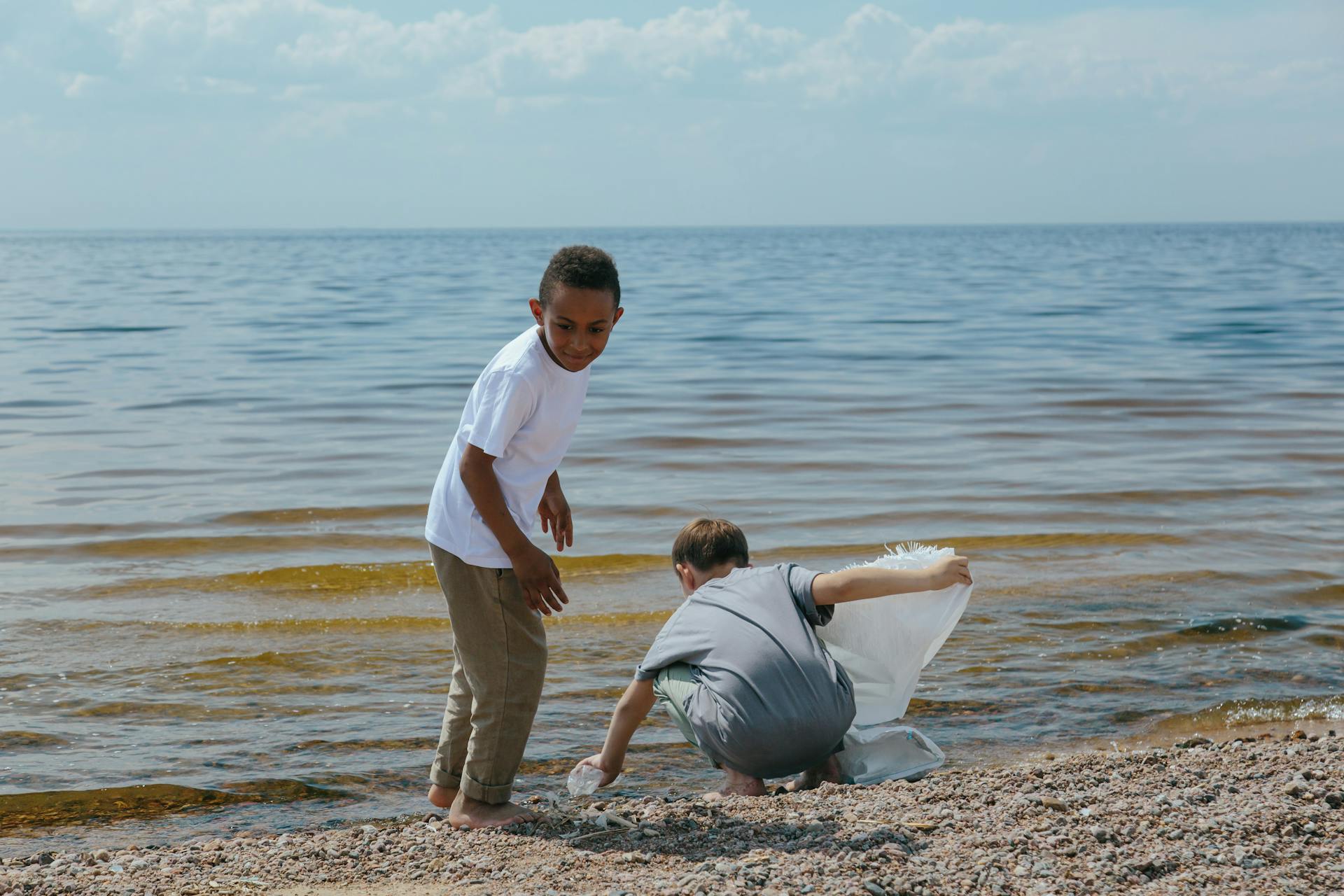 Two children clean up a beach, collecting trash into a bag, promoting environmental awareness.