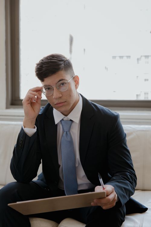 Free Confident young man in suit sitting indoors, adjusting glasses. Stock Photo