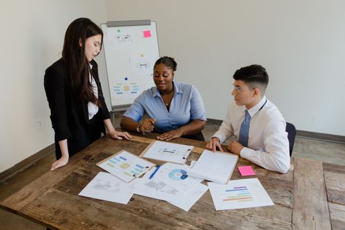 Free Three colleagues discussing work documents at a table, emphasizing teamwork and diversity. Stock Photo
