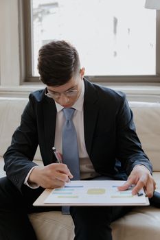 Young businessman in a suit writing on a clipboard while sitting indoors near a window.