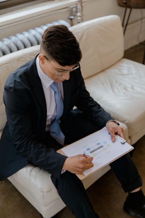 Free A man in a suit reviews documents on a clipboard while sitting on a sofa. Stock Photo
