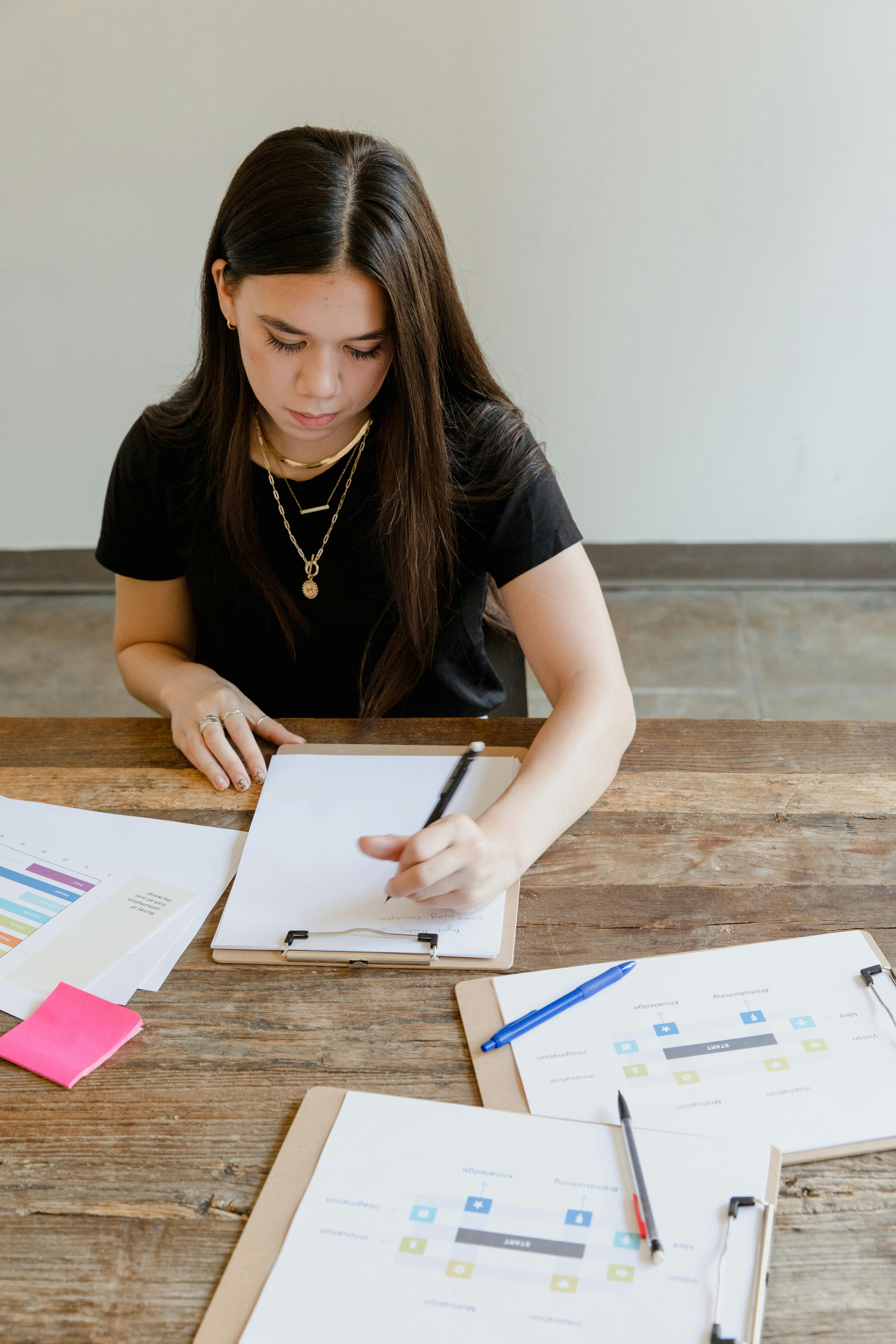 Young woman writing notes at a wooden table in a bright office, focusing on her work.