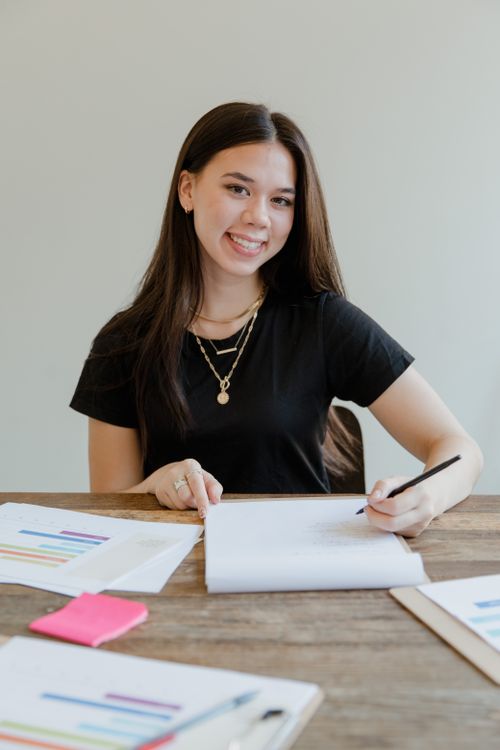 Free Smiling woman in black shirt writing on documents at a wooden desk, indoors. Stock Photo