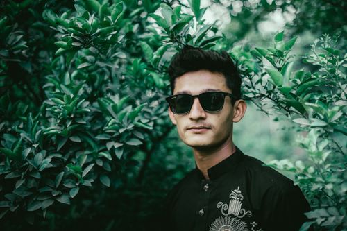 Free Portrait of a stylish man wearing sunglasses surrounded by lush green foliage. Stock Photo