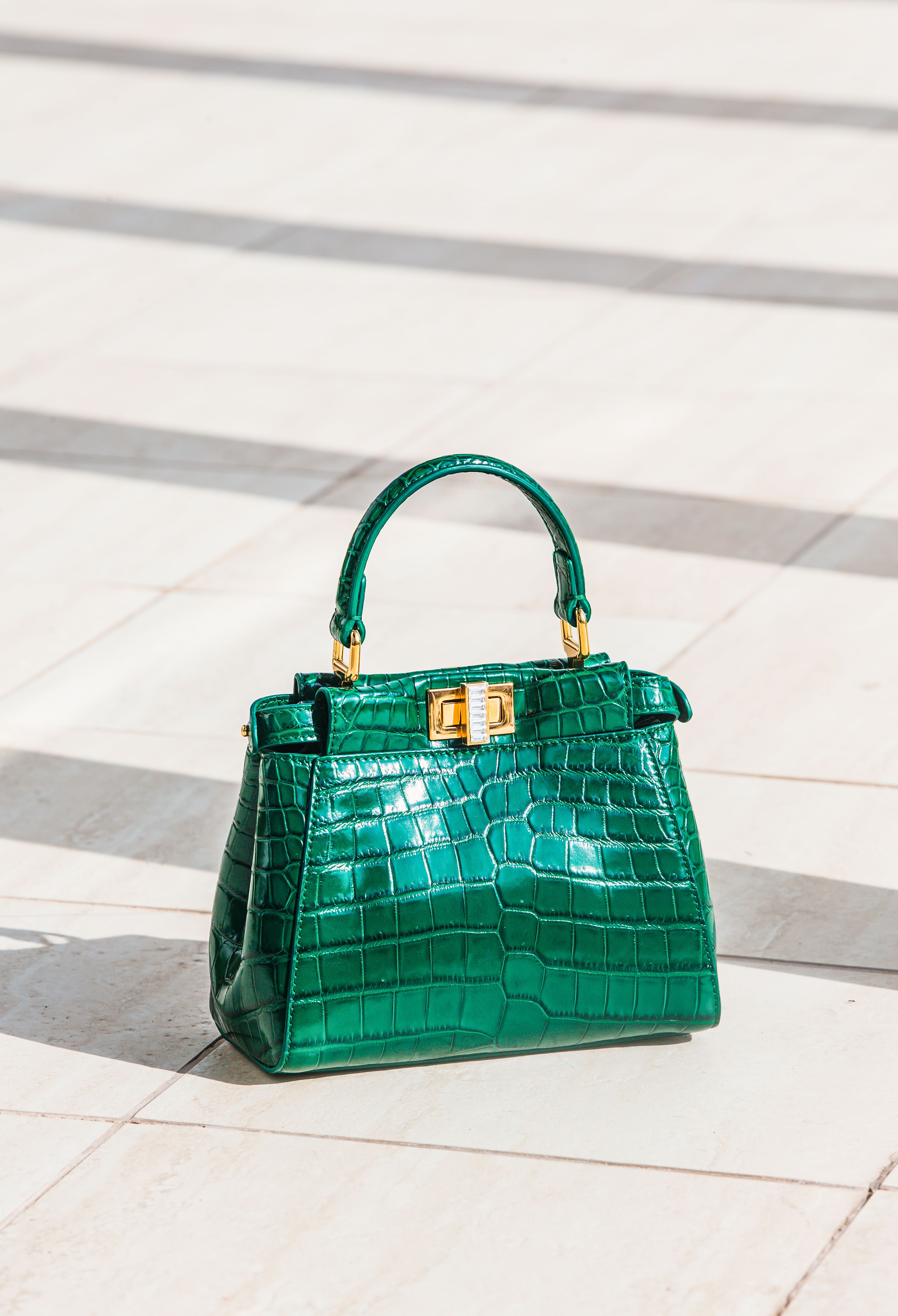 Luxuosa bolsa verde com textura de crocodilo exibida em um pavimento claro ao ar livre, sob luz natural.