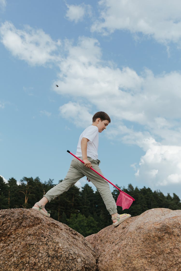 A Boy In White Shirt Walking On Boulders While Holding A Net