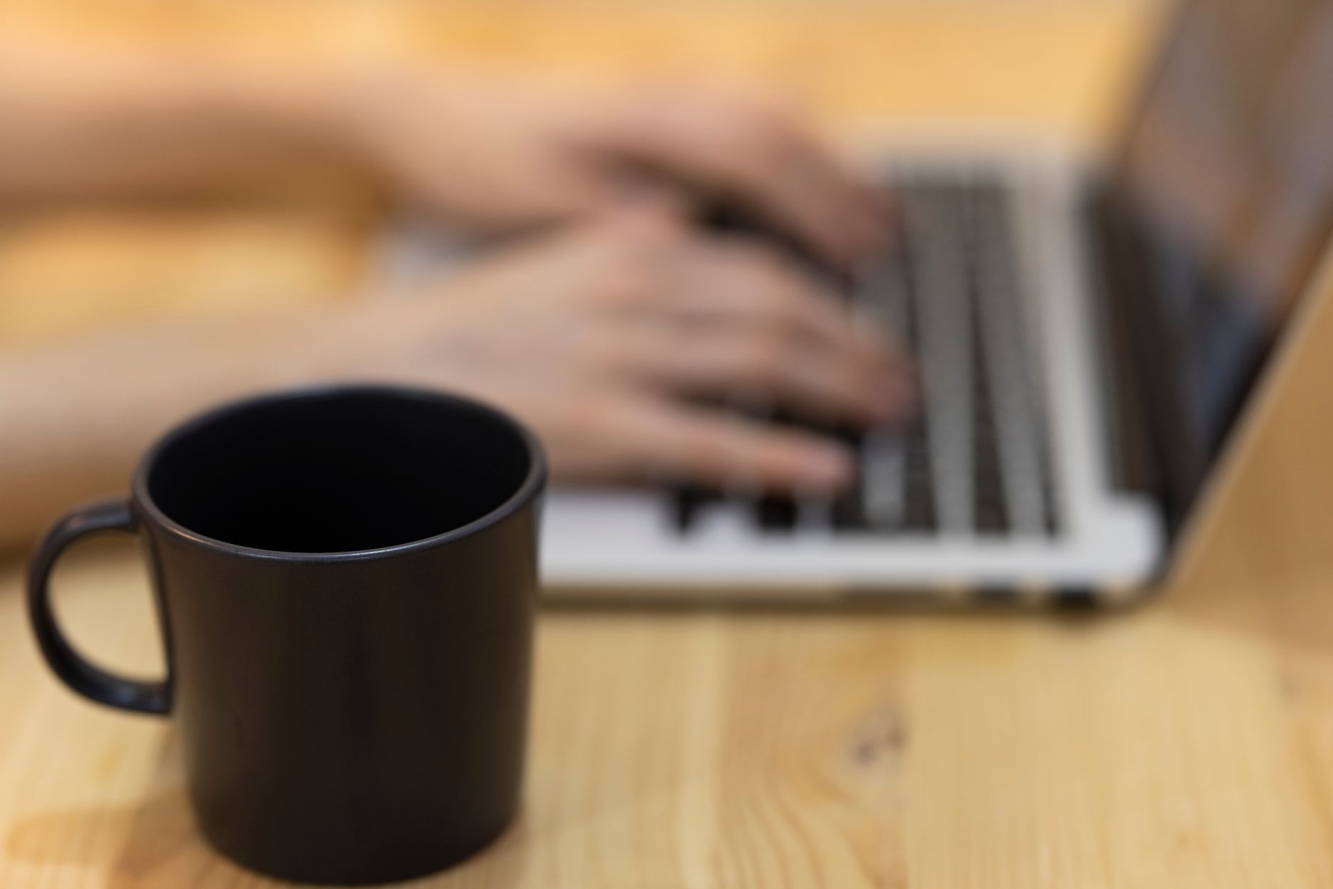 Black mug in focus on wooden table with blurred hands typing on laptop background.