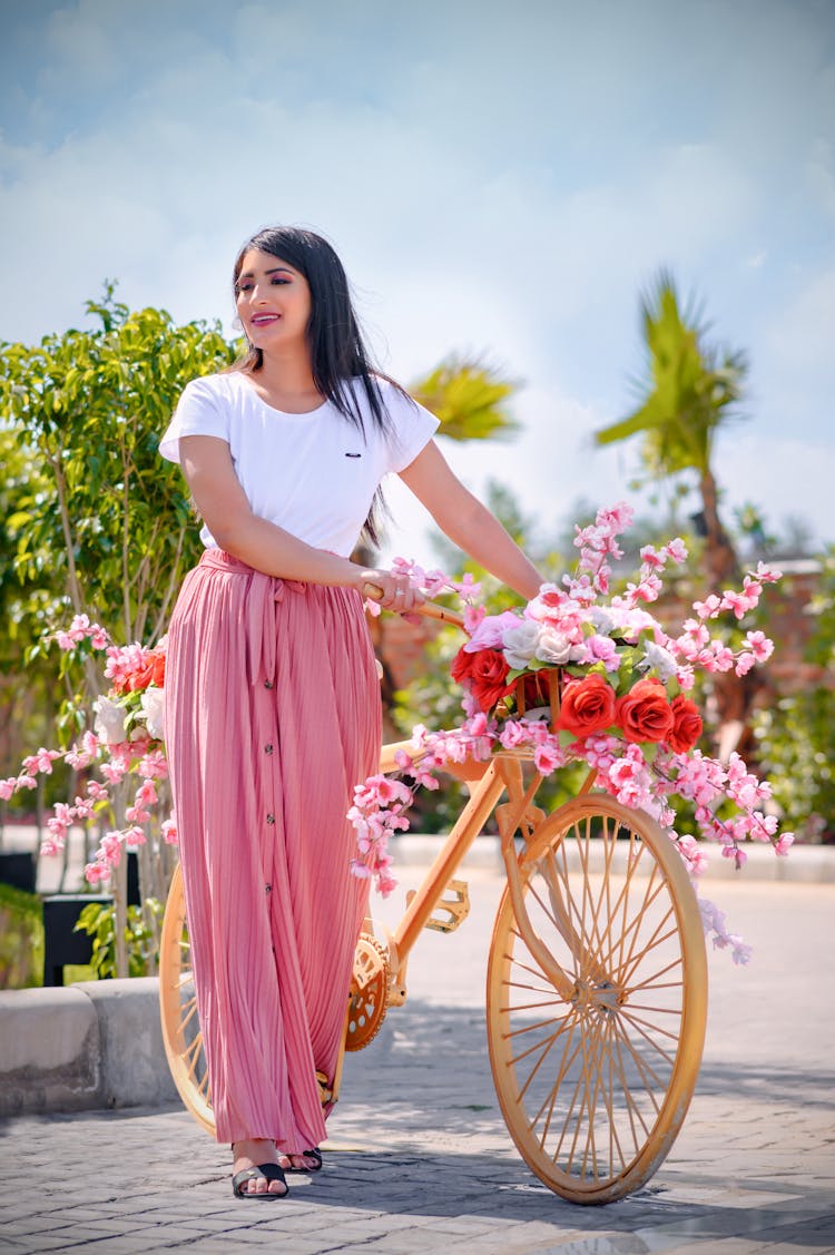 Woman In White Shirt And Pleated Pink Pants Standing Beside A Bicycle With Flowers