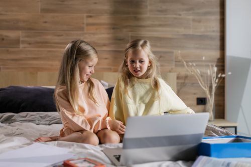 Free Two young girls learning online together at home, sitting on a bed with a laptop. Stock Photo