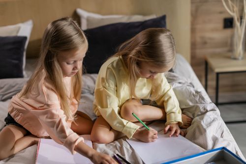 Free Cute twin girls drawing with pencils while sitting on a cozy bed indoors. Stock Photo