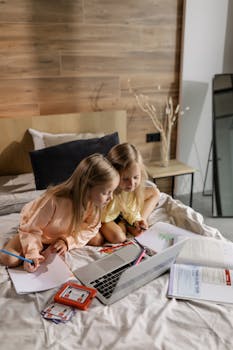 Two young girls studying together on a bed with a laptop, surrounded by books and papers.