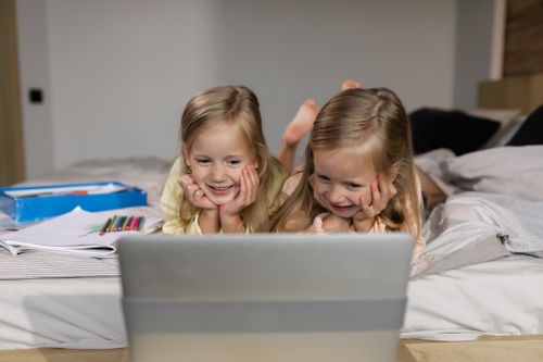 Free Two blonde twin sisters lying in bed, watching a laptop together indoors. Stock Photo