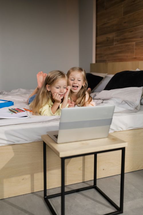 Free Two joyful twin girls learning online, lying on bed with a laptop, exploring digital education at home. Stock Photo