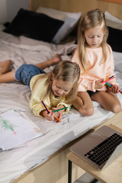 Free Two young girls drawing with colored pencils on a bed while watching a laptop. Perfect for themes of creativity and childhood. Stock Photo