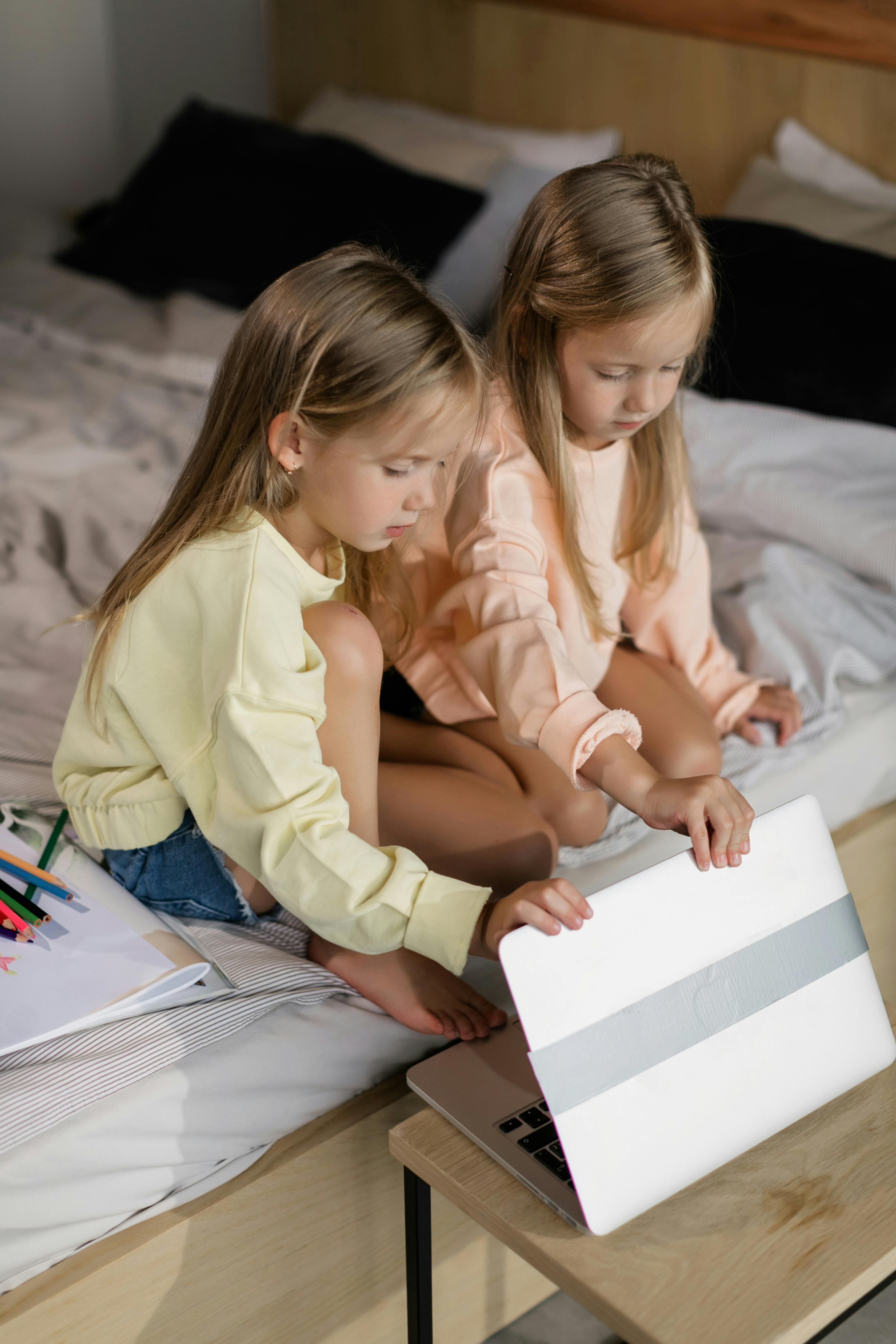 Two young girls sitting on a bed, focused on online learning with a laptop.