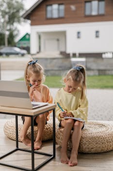 Two young girls learning online together, writing and using a laptop outdoors.