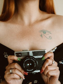 Close-up of a woman holding a vintage camera, showcasing her tattoo and colorful nail art for a creative aesthetic.