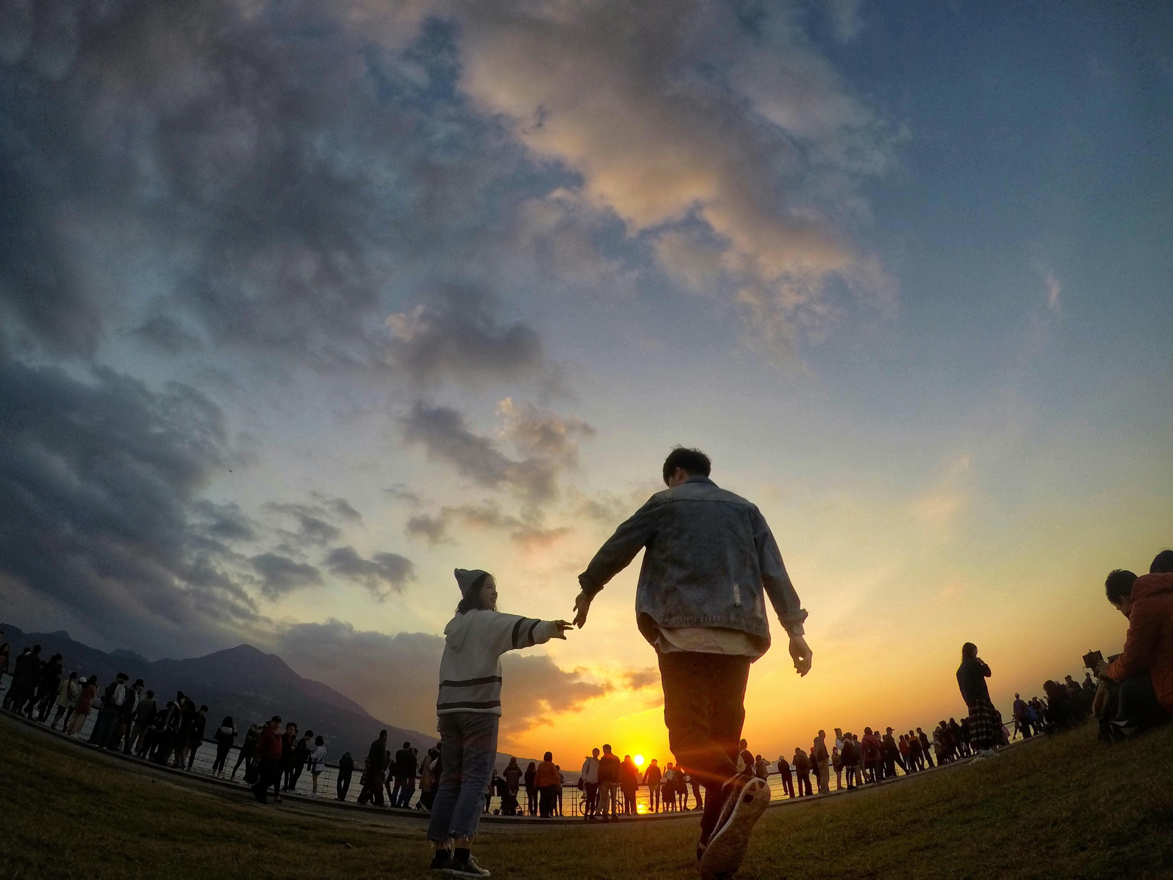 一对夫妇牵着手，在充满活力的日落天空下，背景中映衬着人群
