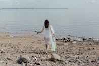 一名穿着透明连衣裙的妇女在平静的海面上拿着一个塑料袋，沿着多岩石的海滩散步