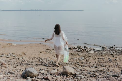 Free A woman in a sheer dress walks along a rocky beach, holding a plastic bag by the calm sea. Stock Photo