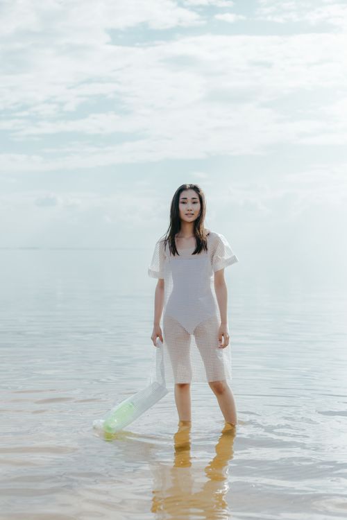 Free A stylish woman stands in shallow water, wearing a see-through dress under cloudy skies. Stock Photo