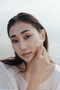 Asian woman gently applying skincare cream to her face outdoors.