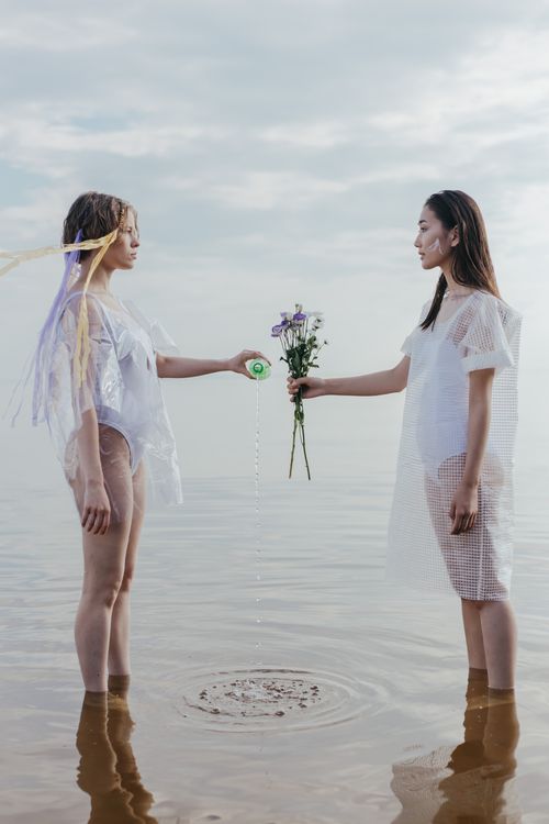 Free Two women standing in water exchanging a water bottle and flowers, creating a serene scene. Stock Photo