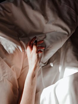 A delicate hand rests on sunlit crumpled sheets, capturing a moment of serene stillness.