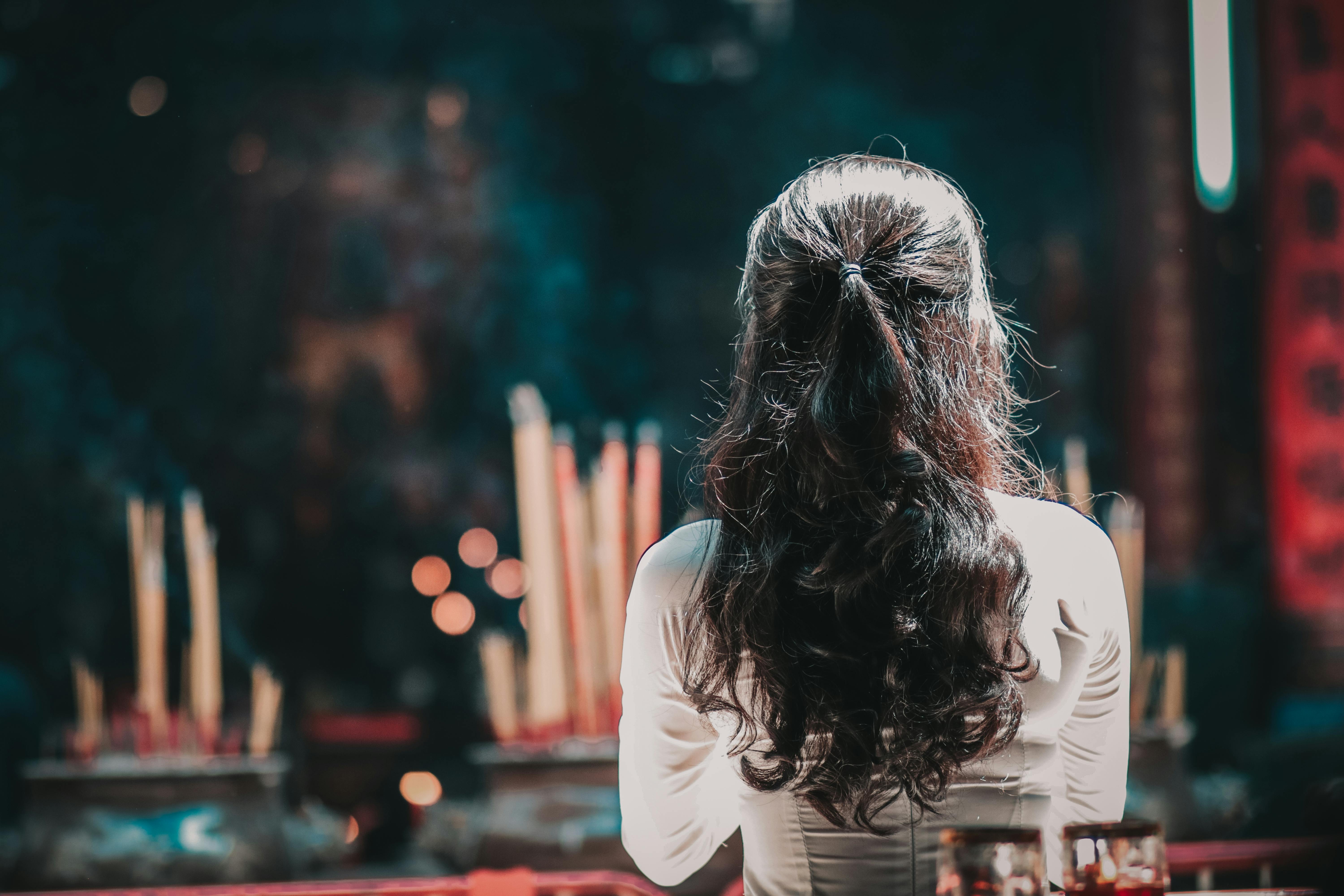 A woman with long hair is seen from behind praying inside a Vietnamese temple, surrounded by incense.
