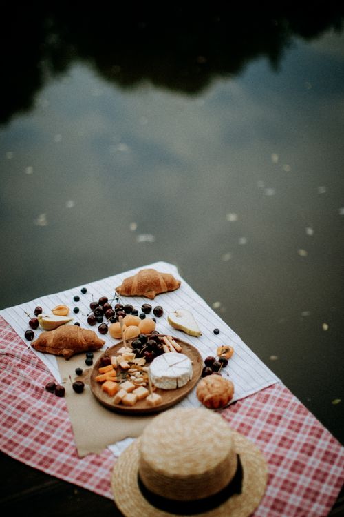 Free Serene picnic scene by the lake with cheese, croissants, and fruits. Stock Photo
