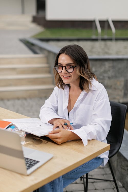 Woman in White Long Sleeve Shirt Sitting at Table with Laptop