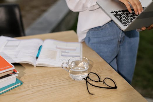 Casual indoor workspace with laptop, open book, eyeglasses, and a cup of water on a wooden table.