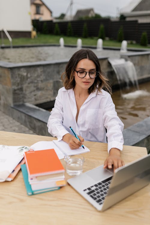 Woman Using a Laptop While Writing on a Notebook