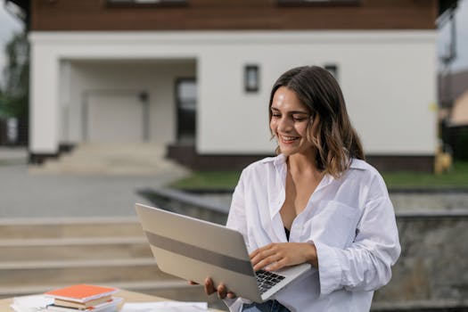 Freelancer smiling while working remotely on laptop outside modern home.