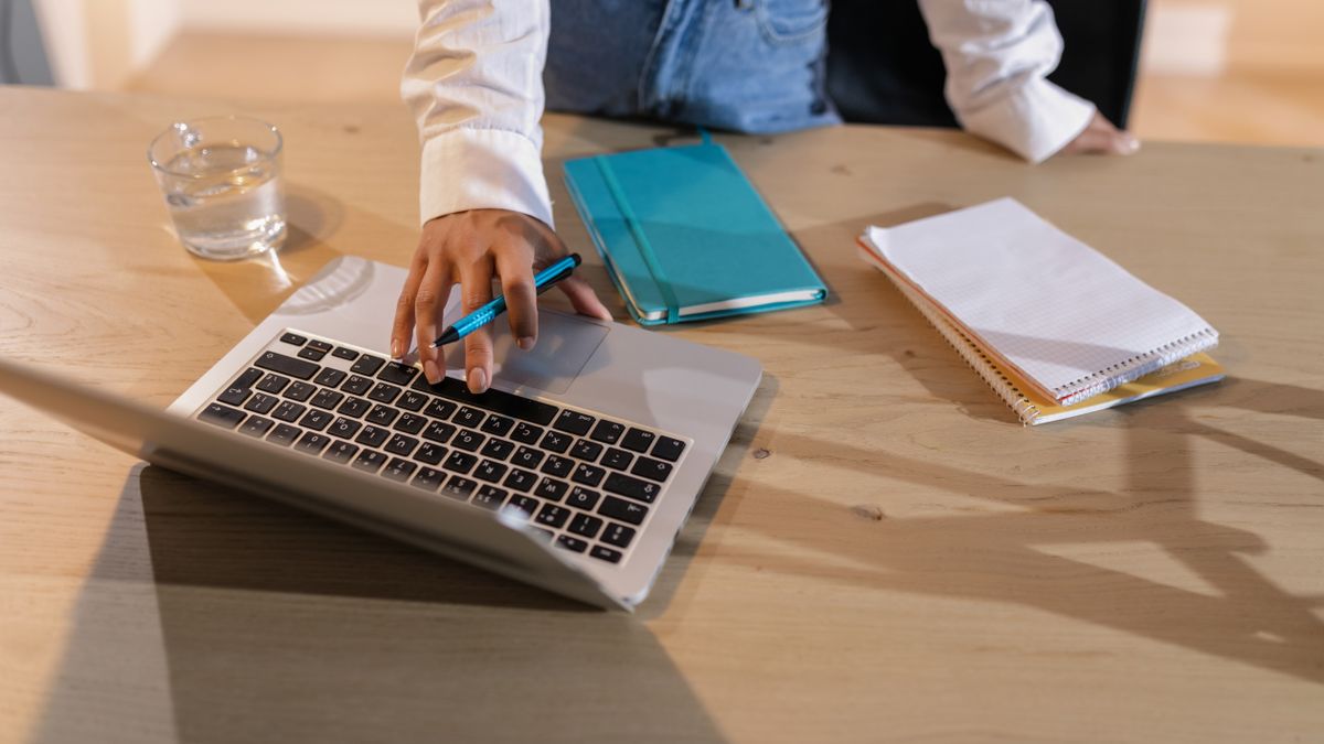 An overhead view of a person using a laptop on a wooden desk with notebooks and a glass of water.