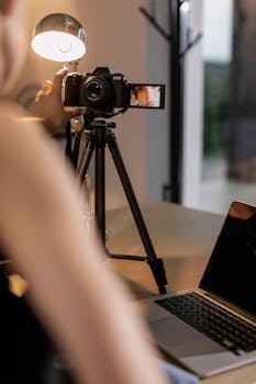 A person sets up a DSLR camera on a tripod for vlogging indoors, illuminated by a lamp.