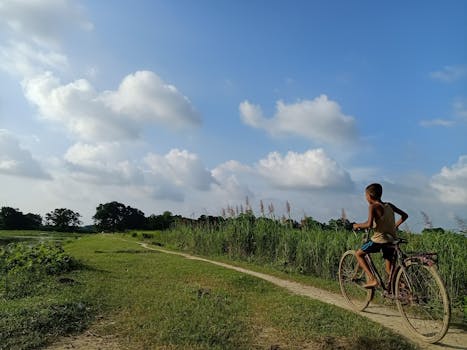 A young boy cycles along a grassy rural path under a bright blue sky with fluffy clouds.