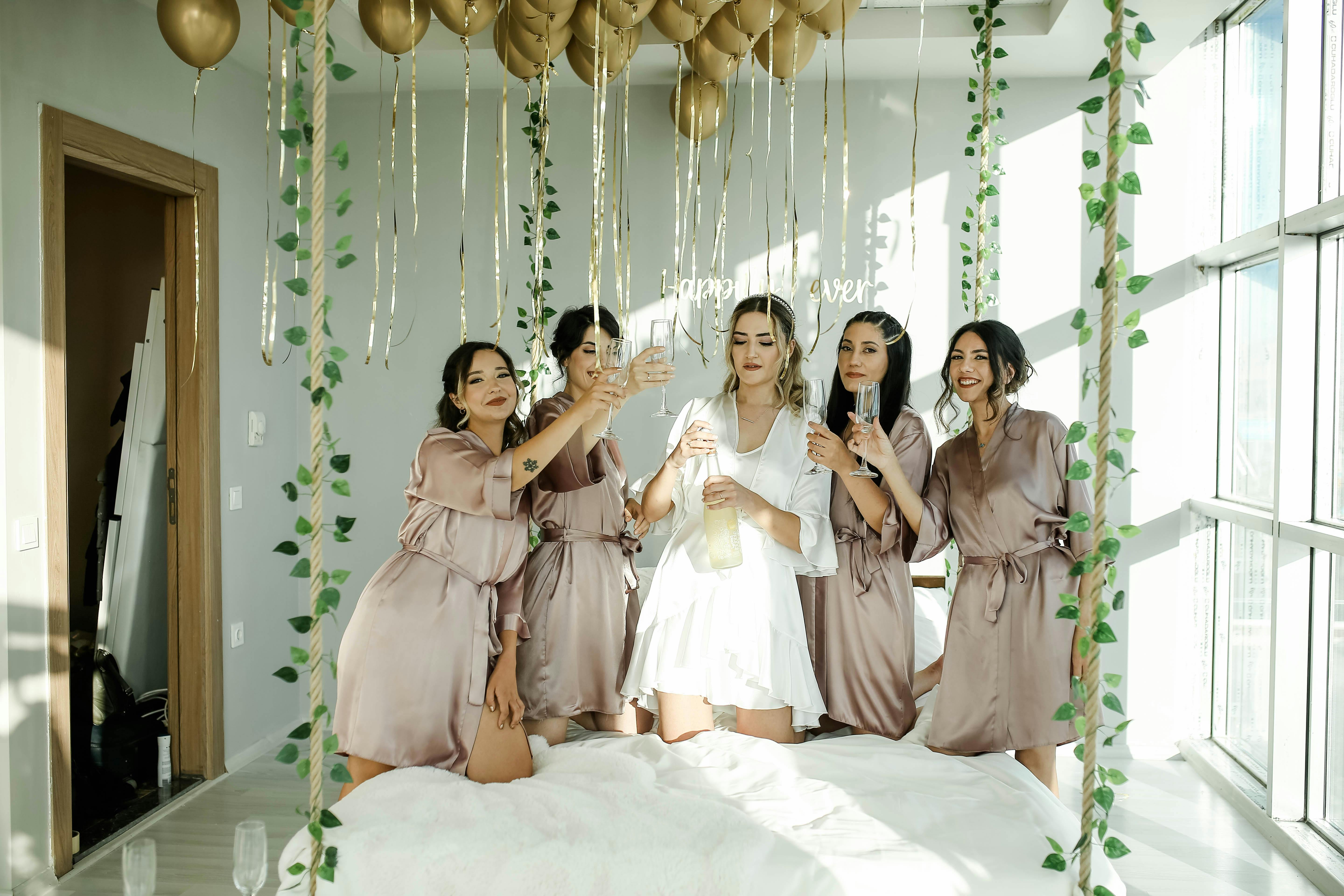 Five women in robes celebrating a bridal party with champagne indoors.