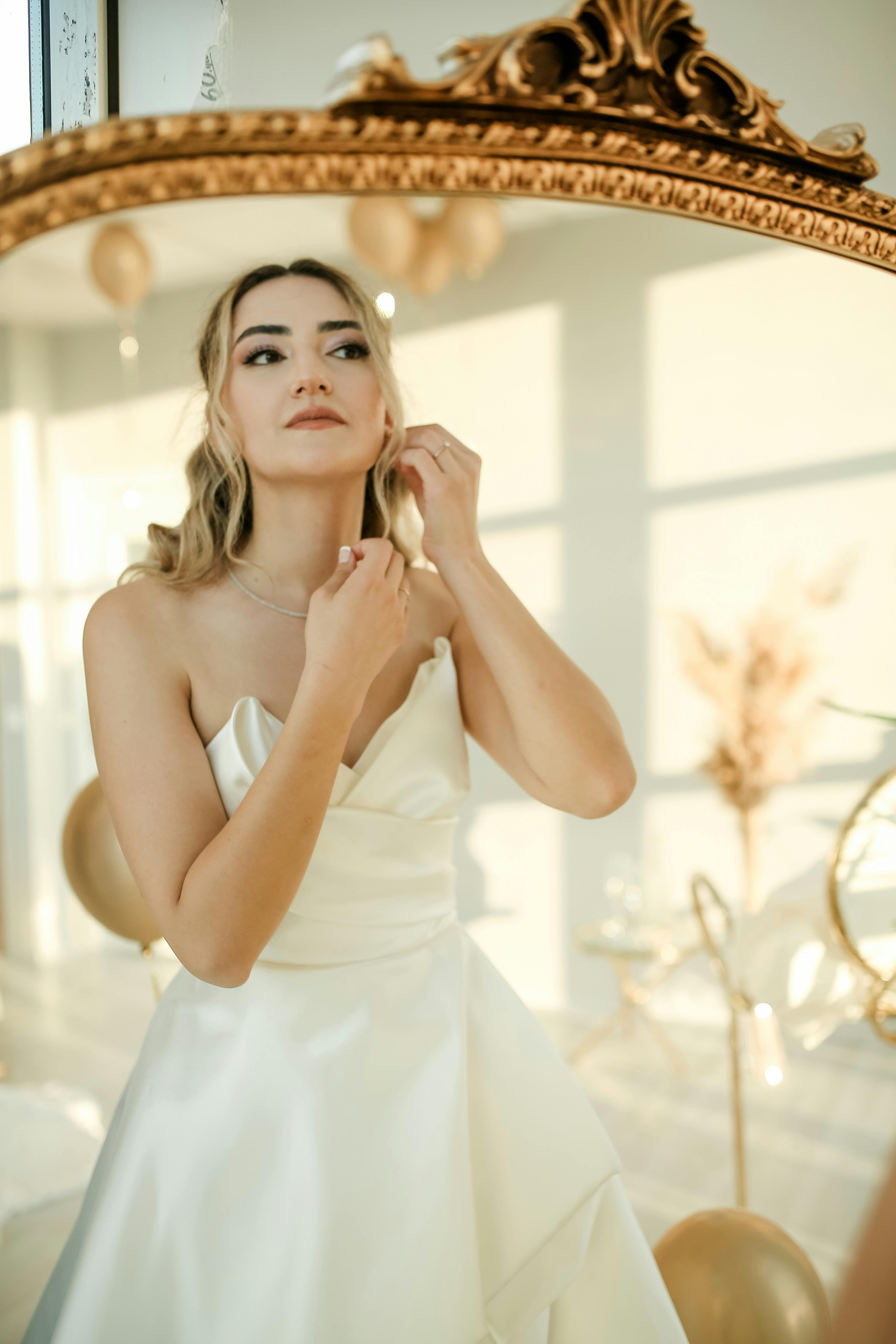 A beautiful bride adjusting her necklace while looking in a decorative mirror, wearing a classic wedding gown.