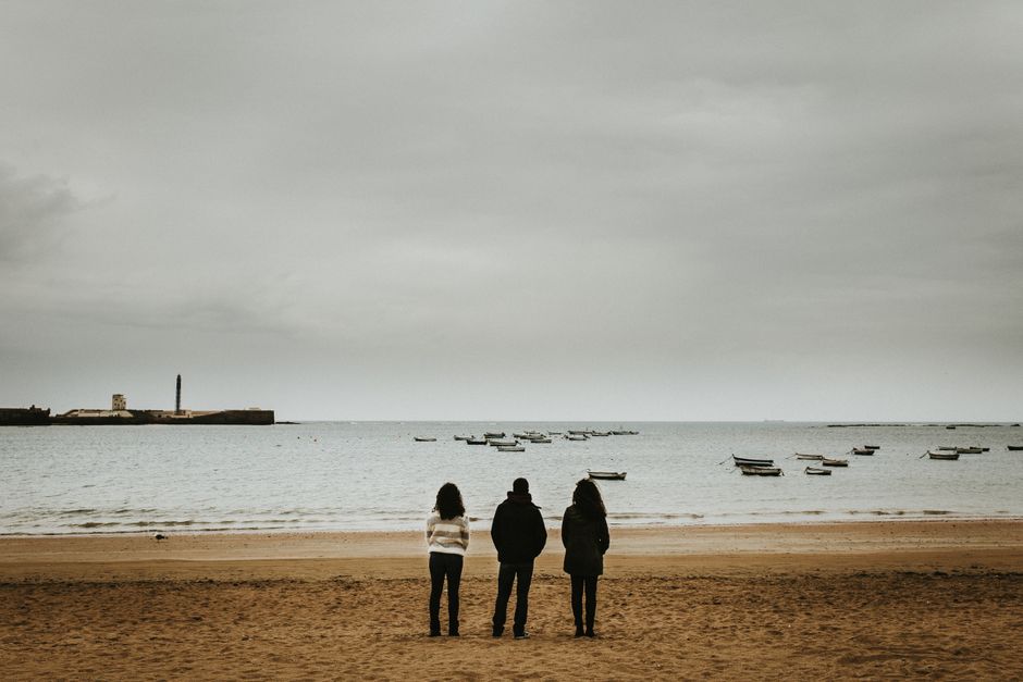 Tres personas de pie en una playa tranquila, contemplando los barcos flotando en el mar.