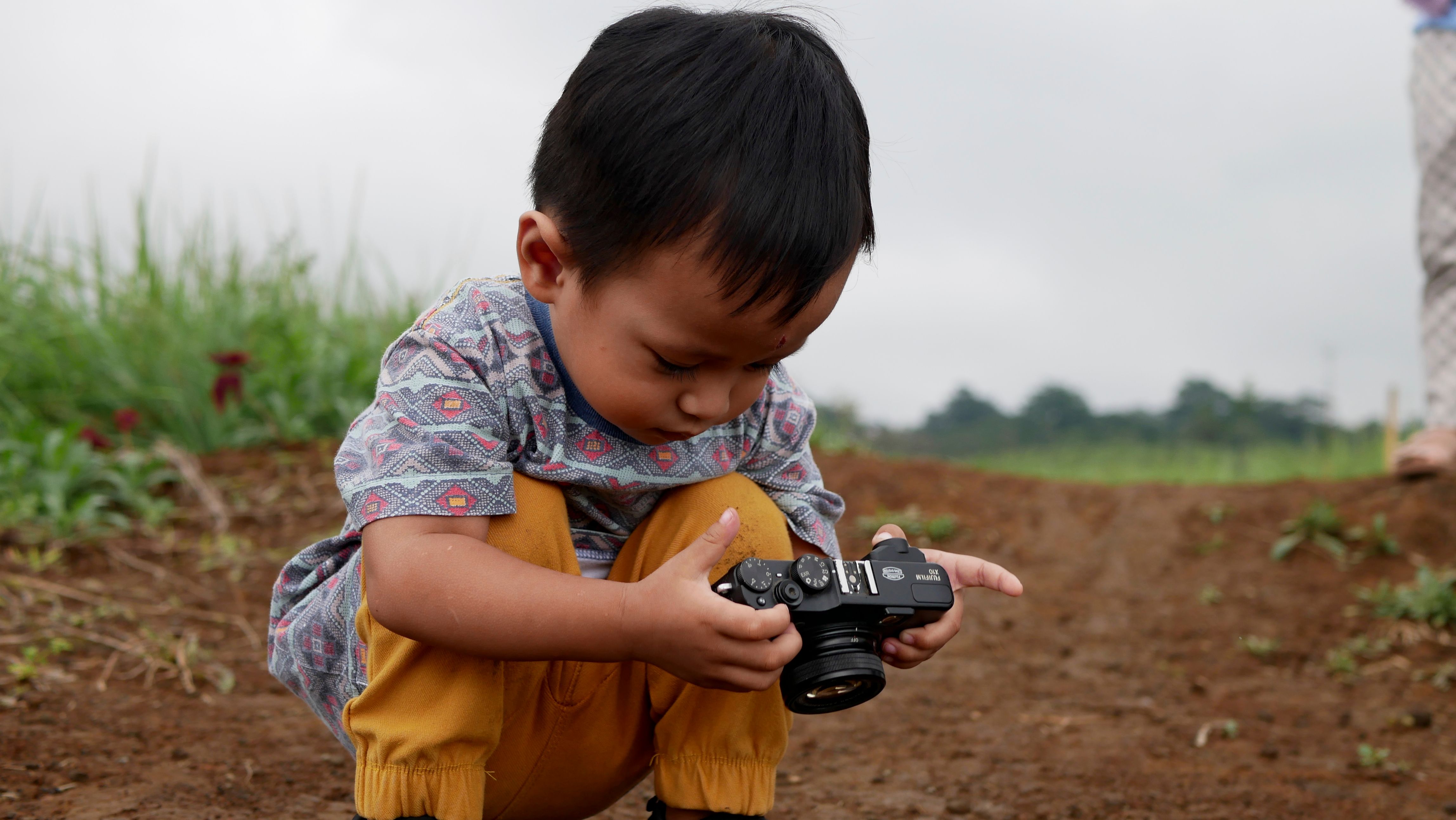 A young child kneels on a dirt path examining a camera outdoors in Jawa Timur, Indonesia.