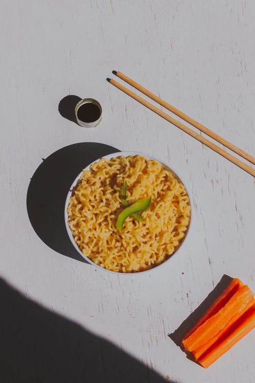 Free Appetizing flatlay photo of noodles garnished with green pepper, carrot sticks, and chopsticks on a sunny white surface. Stock Photo