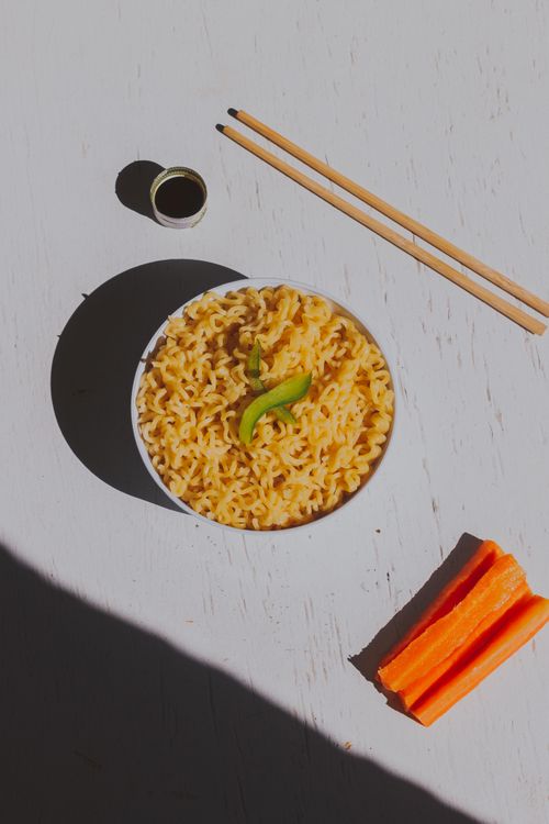 Free Minimalist shot of Asian noodles in a bowl, accompanied by chopsticks and carrot slices on a white background. Stock Photo