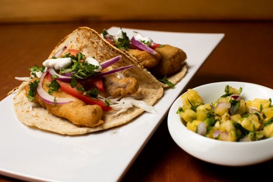 Two battered fish tacos with tomato, red onion, cabbage, cilantro and crema beside pineapple salsa
