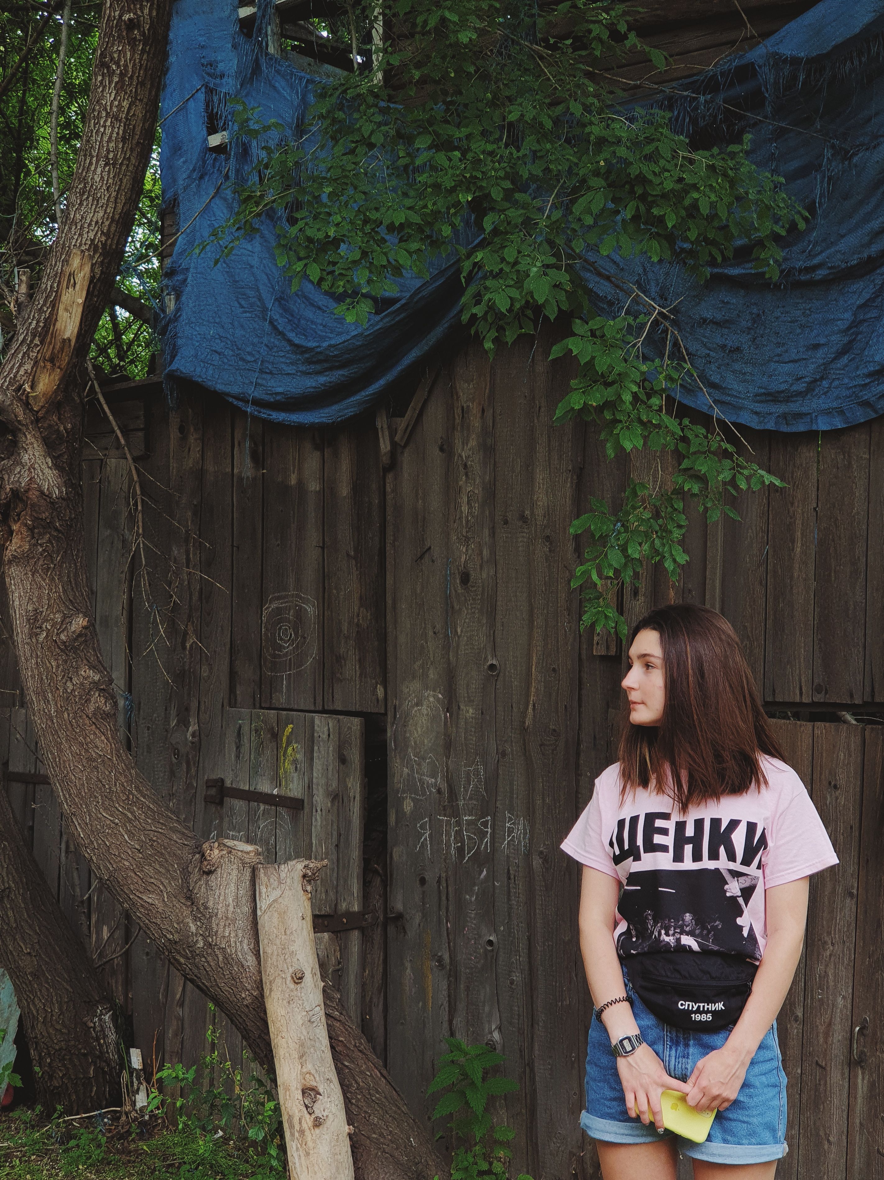 A young woman stands by a rustic wooden wall outdoors, gazing thoughtfully to the side.