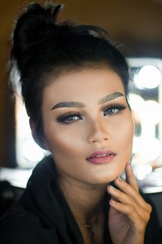 Close-up portrait of a woman with bold makeup and hand on chin, looking directly at the camera.
