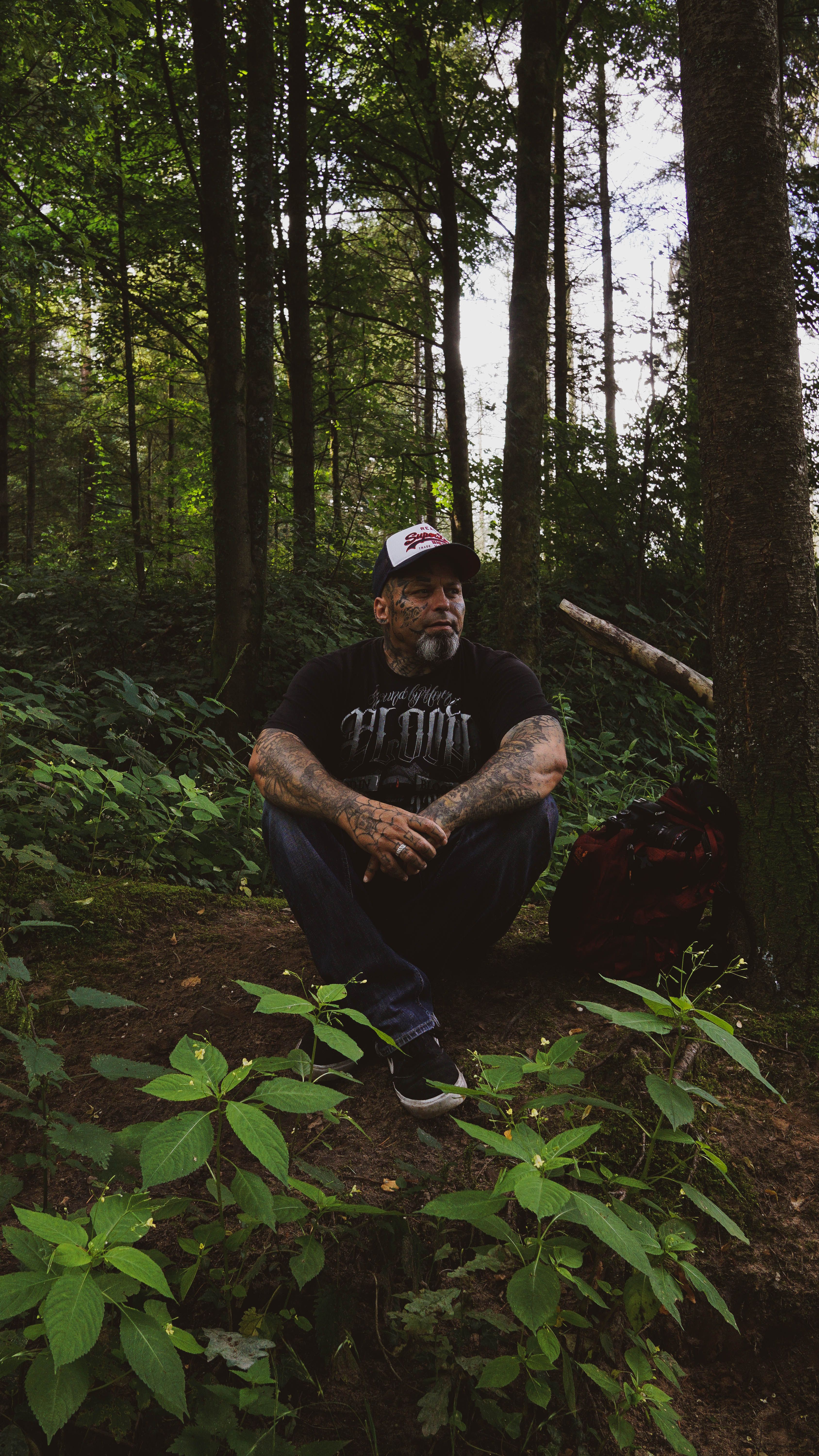 A tattooed man sits peacefully in a lush green forest, surrounded by tall trees and plants.